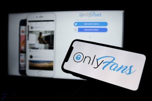 Bilgisayar ekranında OnlyFans'ın ana sayfası, önündeki telefonda OnlyFans'ın logosu görünüyor.