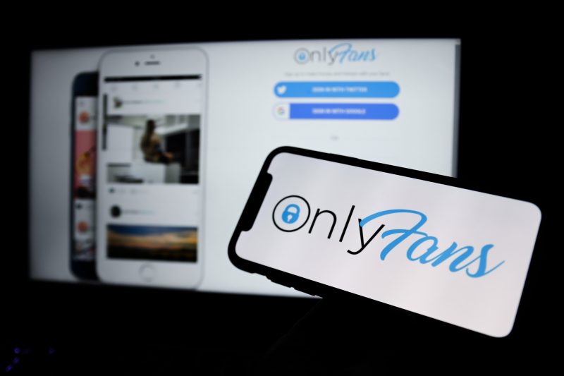 Bilgisayar ekranında OnlyFans'ın ana sayfası, önündeki telefonda OnlyFans'ın logosu görünüyor.