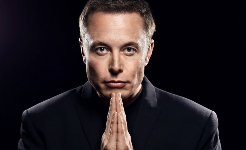 Elon Musk siyah arka plan önünde ellerini çenesinin altında birleştirerek poz veriyor.