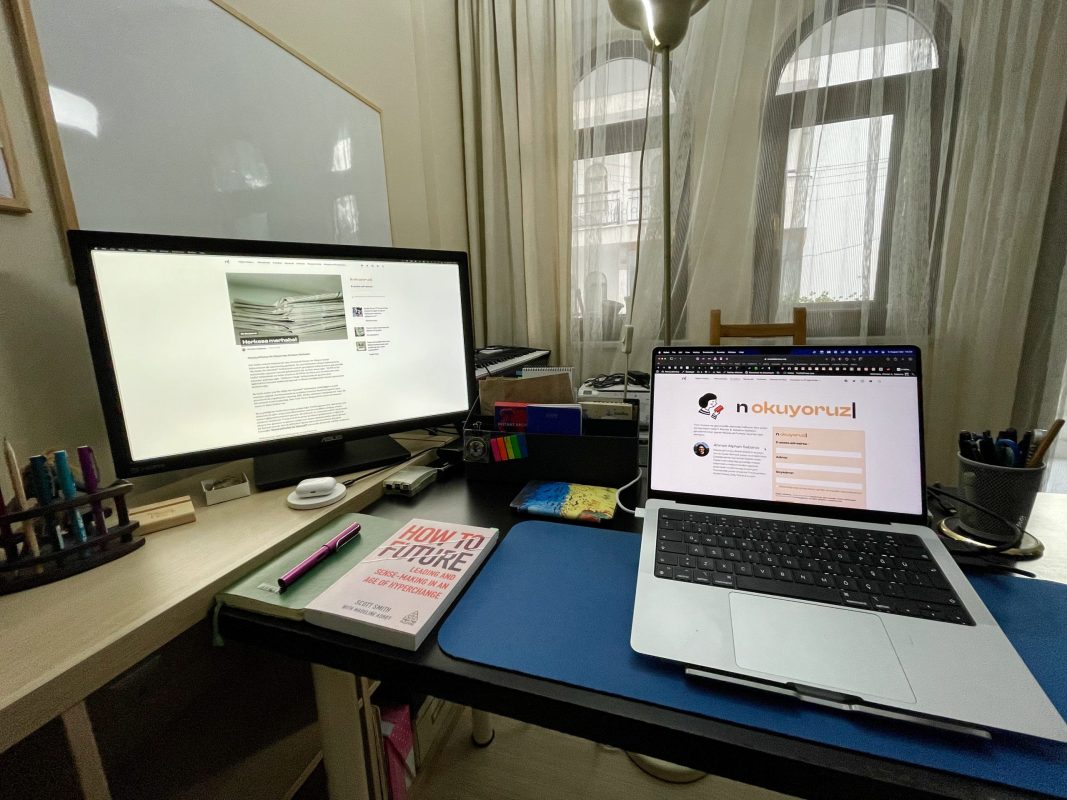 200. bülteni yazdığım çalışma masasının fotoğrafı. Sol tarafta ikinci mönitör, kalemler ve başka ekstralar var. Ön ve sağ taraftaki masada macbook bilgisayar, bir not defteri ve üstünde dolmakalem, yanında How to Future isimli bir kitap, onların arkasına doğru da Kindle, Raspberry Pi gibi ekstra eşyalar duruyor.