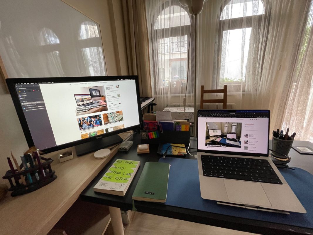 201inci bülteni yazdığım çalışma masasının fotoğrafı. Sol tarafta ikinci mönitör, kalemler ve başka ekstralar var. Ön ve sağ taraftaki masada macbook bilgisayar, bir not defteri ve üstünde dolmakalem, yanında Algoritmalar Ne İster isimli bir kitap, onların arkasına doğru da Kindle, Raspberry Pi gibi ekstra eşyalar duruyor.
