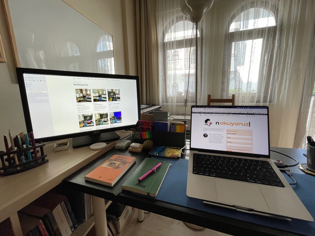 202inci bülteni yazdığım çalışma masasının fotoğrafı. Sol tarafta ikinci mönitör, kalemler ve başka ekstralar var. Ön ve sağ taraftaki masada macbook bilgisayar, bir not defteri ve üstünde dolmakalem, yanında Medya ve Demokrasi isimli bir kitap, onların arkasına doğru da Kindle, Raspberry Pi gibi ekstra eşyalar duruyor.