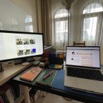 202inci bülteni yazdığım çalışma masasının fotoğrafı. Sol tarafta ikinci mönitör, kalemler ve başka ekstralar var. Ön ve sağ taraftaki masada macbook bilgisayar, bir not defteri ve üstünde dolmakalem, yanında Medya ve Demokrasi isimli bir kitap, onların arkasına doğru da Kindle, Raspberry Pi gibi ekstra eşyalar duruyor.