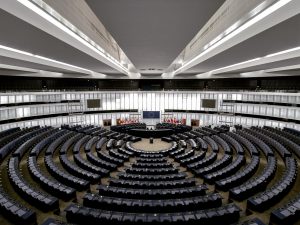 Avrupa Birliği meclisinin boş hâlinin bir fotoğrafı. Tam merkezde ve uzakta AB bayrağı altında kürsüler görünüyor, etrafı ise boydan boya koltuklarla çevrili.