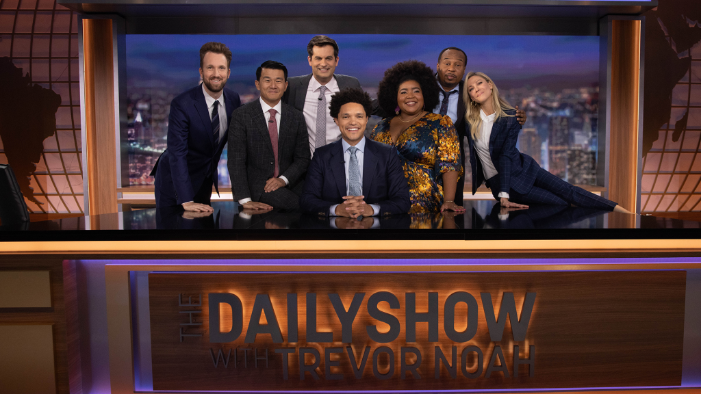 The Daily Show'un sunucusu Trevor Noah, önünde programın adı yazan masasında oturuyor, arkasında ise onunla birlikte programda yer alan altı ekip arkadaşı duruyor ve hepsi gülümseyerek poz veriyorlar.