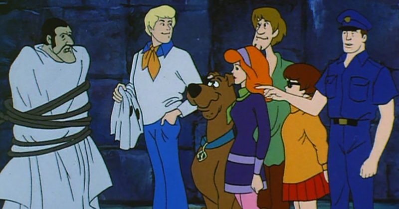 Scooby Doo ekibi bağlayıp maskesini çıkardıkları bir kötü karakterin başında duruyor ve yanlarındaki polise bir şeyler anlatıyor.