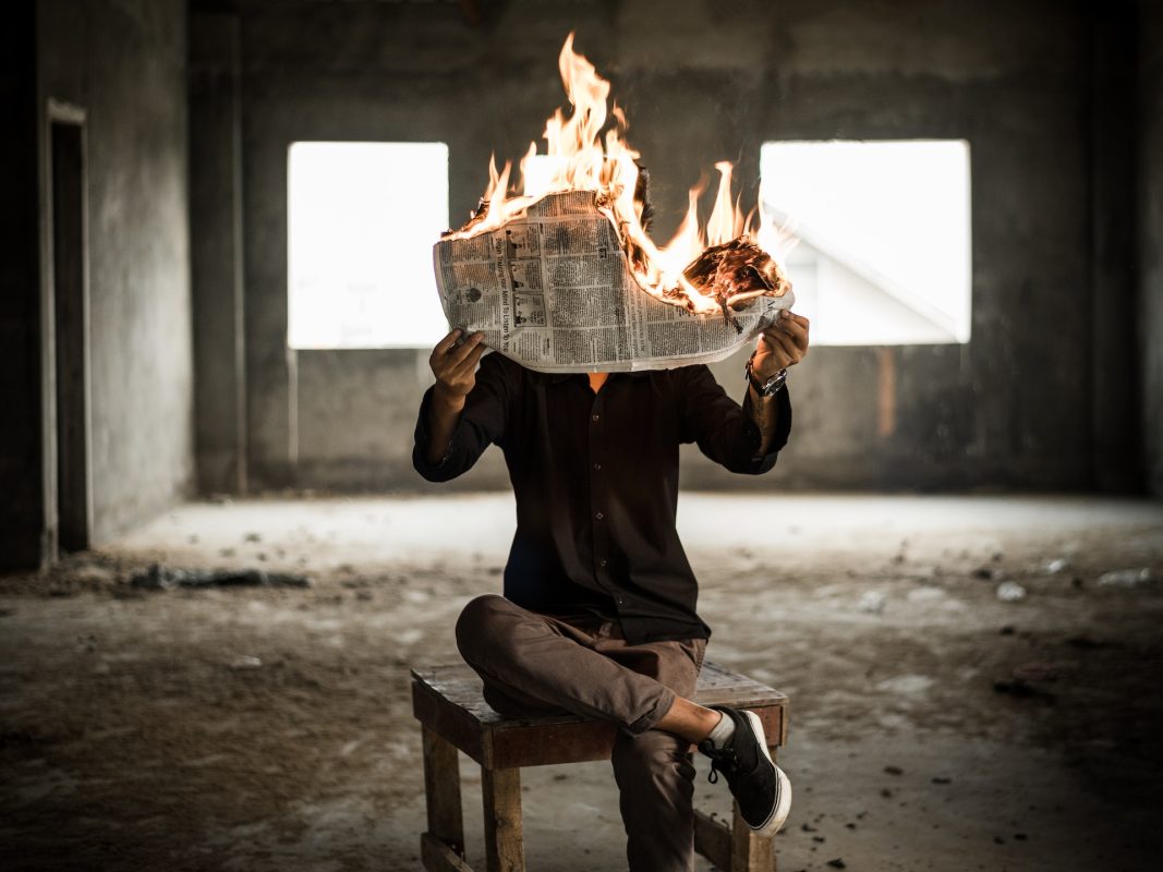 Terk edilmiş bir binanın içerisinde oturan birisi elindeki alev almış gazeteyi okuyor.