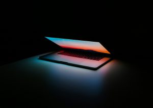 Karanlık bir odanın içerisinde yarım açık şekilde duran ve ekranı parlayan bir Macbook.
