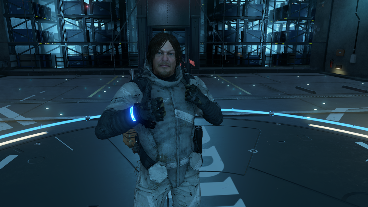 Death Stranding oyunundan aldığım bu ekran görüntüsünde ana karakter bir deponun içerisinde tuhaf bir surat ifadesi ve bir elinin baş parmağını yukarıda tutarak kameraya poz veriyor.